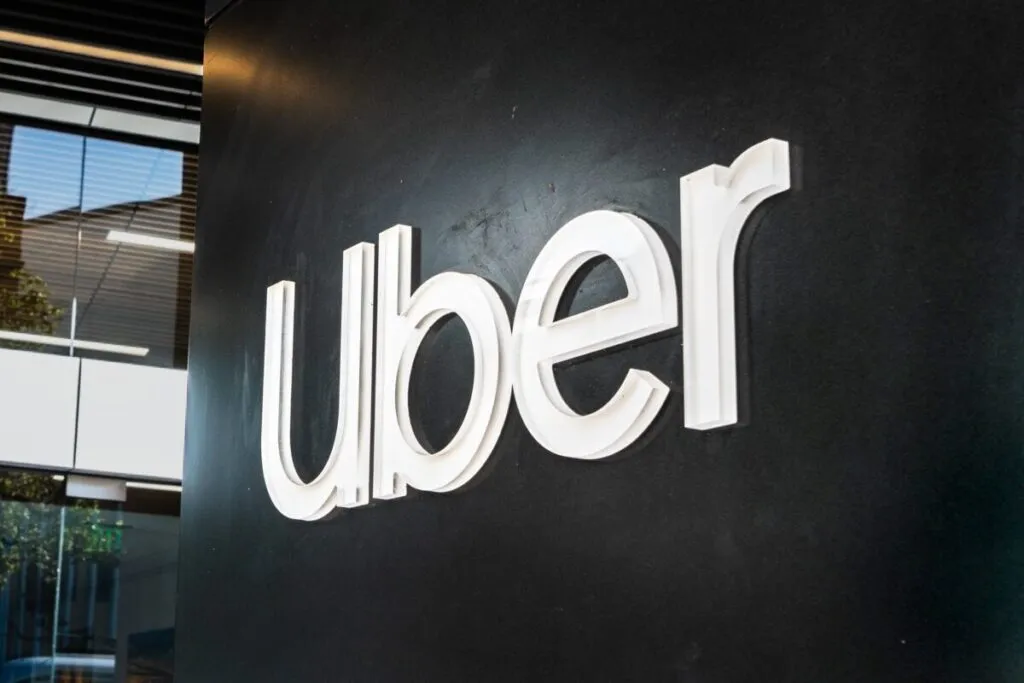 Logotipo tridimensional de Uber en una pared negra dentro de las oficinas de la compañía, con reflejos de luz y parte del entorno moderno visible a través de ventanales de vidrio.
