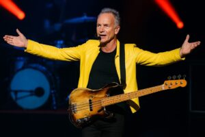 Sting con chaqueta amarilla tocando el bajo sobre el escenario, en concierto reciente en Florida antes del Super Bowl